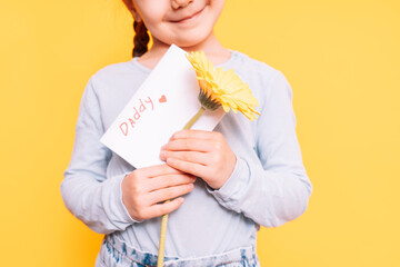 Child holding drawn card and flower for Father's Day holiday in front of yellow background
