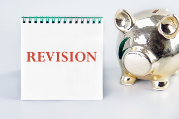 Revision inscription on a vertically standing notebook near the piggy bank on a light background