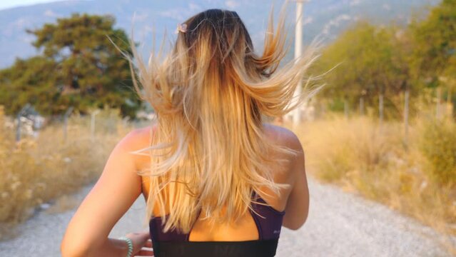 Young blonde girl jogging along country road. Unrecognizable sporty woman working out outdoor. Female sportsman doing run training. Concept of healthy active lifestyle. Slow motion Back view Close up