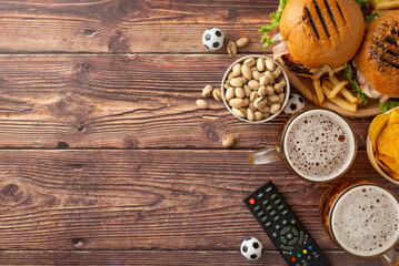 Football frenzy fare. Top view of snack assortment: crispy tortilla chips, savory pistachios, hamburgers, ice-cold beer, soccer balls, remote control, on wooden table with empty space for text