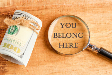 Diversity, business, inclusion and belonging concept. You belong here symbol through a magnifying glass, they are visible on a beautiful background next to a roll of paper banknotes