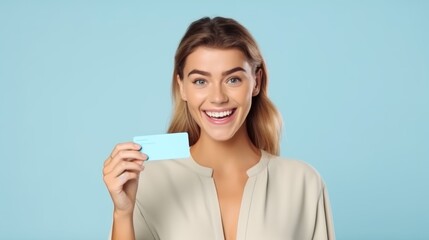 Cheerful young blonde woman holding a credit card, smiling broadly against a soft blue background