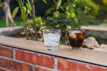 2 glasses filled with ice, 1 with water and 1 with soft drink, on a brick wallflower outdoors on a...