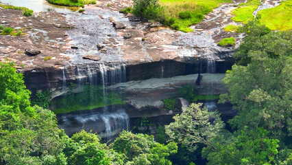 Enchanting Torrent: The waterfall, a marvel, cascades gracefully down a lofty cliff, ensconced within the embrace of a vibrant, tropical woodland. Pha Taem National Park, Thailand.
