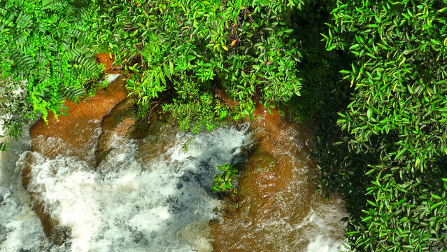 A dense green rainforest conceals a jewel - a waterfall gracefully cascading down layered rocks. A spectacle from above, revealed by a drone's keen eye. Exotic Rainforest: Rich Biodiversity. Thailand. - Powered by Adobe