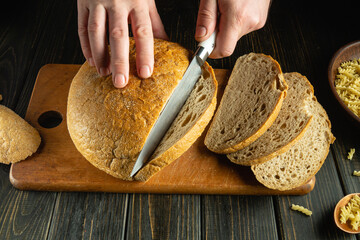 Chef hands cutting fresh bread with a knife on a kitchen board before dinner. Slicing rye bread on...