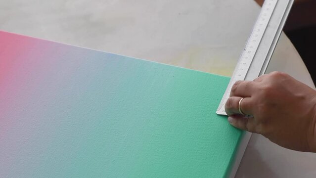 A diverse biracial female artist, measures out a border with a metal ruler on canvas. The canvas has soft pastel gradient background