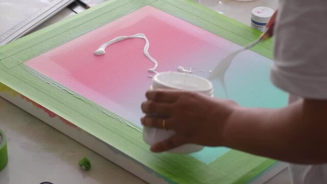 A diverse biracial female artist, paints  a white rectangle using masking tape, paint brush and palette knife on a gradient painted artist canvas.