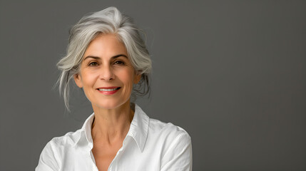 Fototapeta premium Stylish senior woman with grey hair in a white shirt smiling and looking at the camera , grey background
