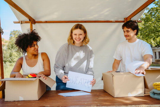 Portrait of happy volunteer holding name tag with male and female colleagues near table during charity drive at communit