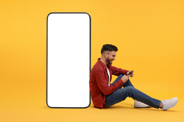Man with mobile phone sitting near huge device with empty screen on orange background. Mockup for design