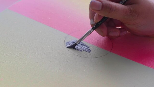 A diverse biracial female artist, paints a grey circle onto a painted artist canvas. The canvas has soft pastel gradient background