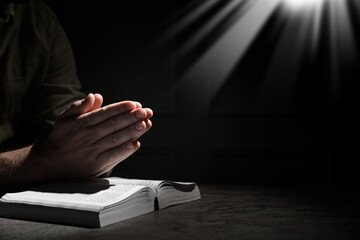 Christian man praying over Bible in darkness, closeup. Holy light shining at him