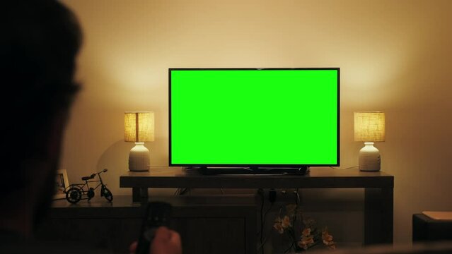 Person watching television with chroma green screen. Unrecognizable female person switching channels on tv with remote controller, sitting on sofa in living room.
