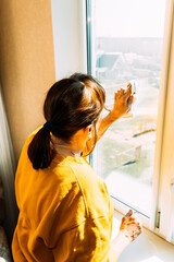 Woman Of Fifty In Yellow Sweater And Jeans Washes Dusty Window In Apartment. 50 Year Old Woman Cleans Windows From Stains Using Rag And Spray Cleaner. Caucasian Elderly Woman Is Cleaning House, Doing