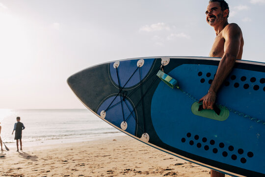 Side view of smiling shirtless man with skateboard standing on sand at beach