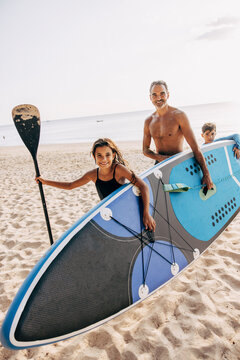 Shirtless man carrying surfboard with daughter and son while walking on sand at beach