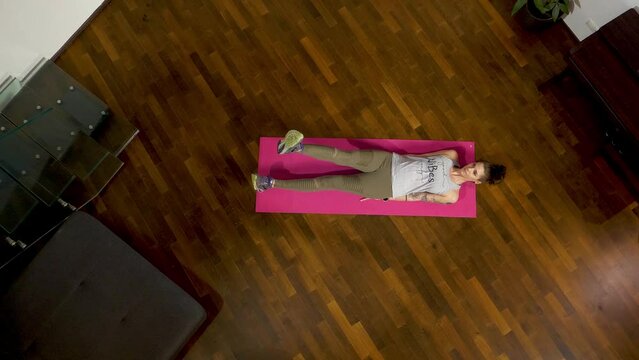 Woman doing flutter kicks on a pink mat, hardwood floor, top-down view, with a tattoo