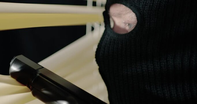 Extreme close-up of criminal with gun and ski mask on looking out window at night.