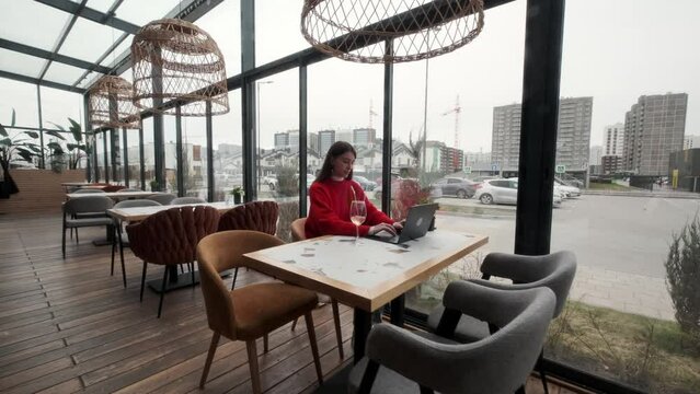 A beautiful young girl in a red jacket is sitting at a table in a restaurant. Working at a laptop. Remote work concept.