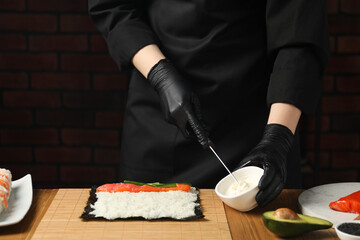 Chef in gloves putting cream cheese onto unwrapped sushi roll at wooden table, closeup