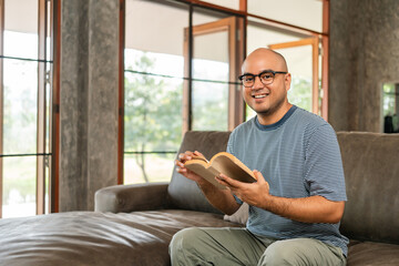 Asian man sitting reading book on cozey couch sofa in living room with sun light morning. People...