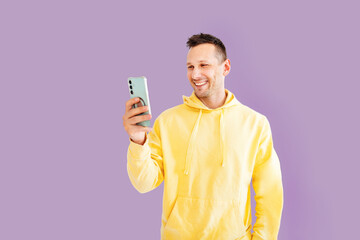 Young smiling happy cheerful Caucasian man 30s in yellow hoodie doing selfie shot on mobile cell phone post photo on social network isolated on pink background studio portrait.