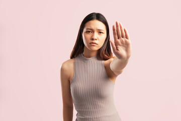 A young Asian woman making a stop sign with her hand, wearing a nude brown shirt on a pink background with empty copy space for advertising content