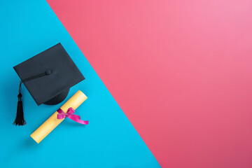 Black graduation cap with a tassel lying next to a rolled yellow diploma tied with a pink ribbon, displayed against a bright blue and pink dual-color background