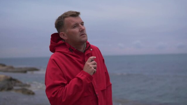 A man in a bright red jacket smokes an electronic cigarette in the evening by the sea alone