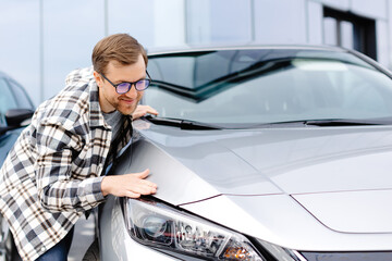 Young bearded man embracing his new car at the dealership smiling joyfully
