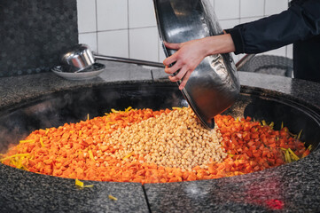 male chef adds chickpeas to cauldron with carrots to cook traditional oriental Uzbek pilaf in...