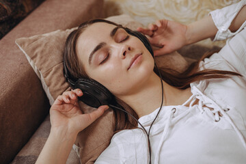 Peaceful woman with closed eyes listening to calm music in headphones while lying on bed at home 