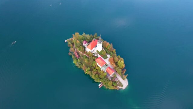 Bird's Eye view of a colourful autumn sunrise over Church of the Assumption of Maria at Lake Bled with athletes rowing in a quad scull across a sunny lake. The sun is reflected on the water, Slovenia