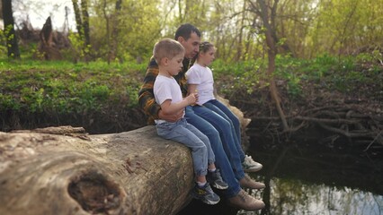 dad with children sits on a log. happy family child dream concept. happy young dad sitting with his son and daughter in the forest on a large log above the river. kids lifestyle dad's