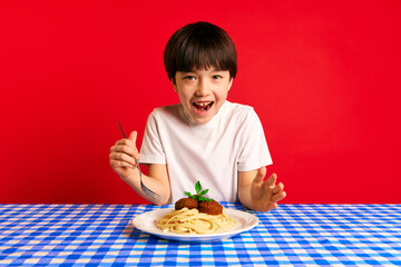 Young boy feeling excited, sitting at table with blue checkered tablecloth and plate of spaghetti and meatballs against red background. Concept of food, childhood, emotions, meal, menu, pop art