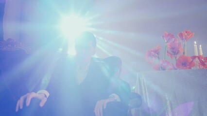 a man in a suit is sitting in a chair in front of a table with candles. A confident guy sits airily on a chair in the rays of artificial light at a party or social event.