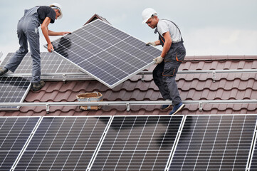 Men technicians carrying photovoltaic solar moduls on roof of house. Electricians in helmets...