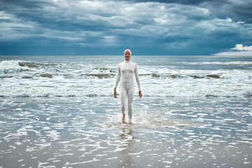 Brave hairless girl with alopecia in white futuristic suit comes out of cold sea on sandy beach,...