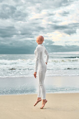Candid hairless girl with alopecia in white futuristic suit twirls in jump on sea sandy beach,...