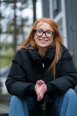 Portrait of a woman. The woman is sitting. The woman is wearing blue jeans and a black windbreaker. The woman has red hair. The woman has a camera.