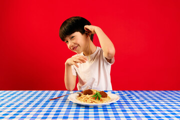 Little smiling boy sitting at table with pasta and meatballs, putting spaghetti on ear against red...
