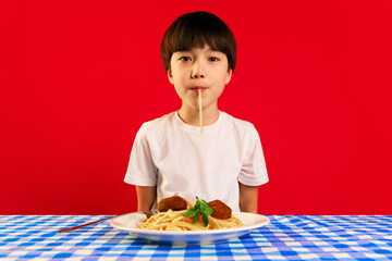 Korean boy, child in white t-shirt sitting at table and eating spaghetti with meatballs against red background. Enjoying delicious dinner. Concept of food, childhood, emotions, meal, menu, pop art