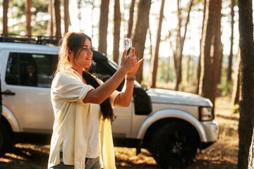 Beautiful woman is standing in the forest with smartphone in hands. Against modern car
