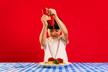 Smiling child, little boy sitting at table and squeezing ketchup sauce on plate with pasta and meatballs against red background. Concept of food, childhood, emotions, meal, menu, pop art