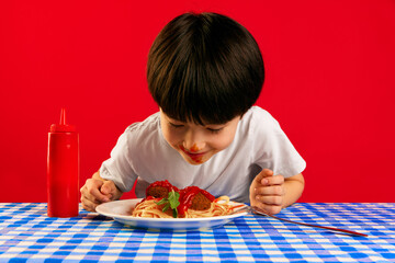 Little boy, child with dirty ketchup face sitting at table and eating delicious spaghetti with meatball and tomato sauce against red background. Food, childhood, emotions, meal, menu, pop art concept