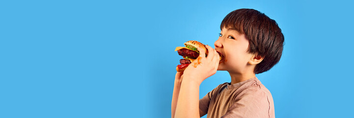 Little boy, kid biting delicious giant burger with meat, vegetables and cheese against blue...