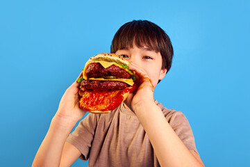 Little boy, kid holding giant, juicy fresh burger with meat, tomato sauce and cheese, eating with delightful face against blue background. Concept of food, childhood, emotions, meal, menu, pop art