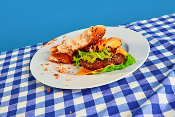 Half-eaten cheeseburger with lettuce, meat, bun and tomato lying on plate on checkered tablecloth...