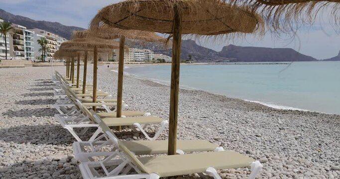 Footage of the beautiful beach in Altea, Alicante in Spain showing sun loungers and straw beach umbrellas on the pebble beach known as Playa La Roda